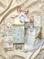 From the Chateau Mail Club collection featuring a monthly letter, château-inspired paper goods, miniature room craft, artwork, tags, and stationery arranged on ivory satin